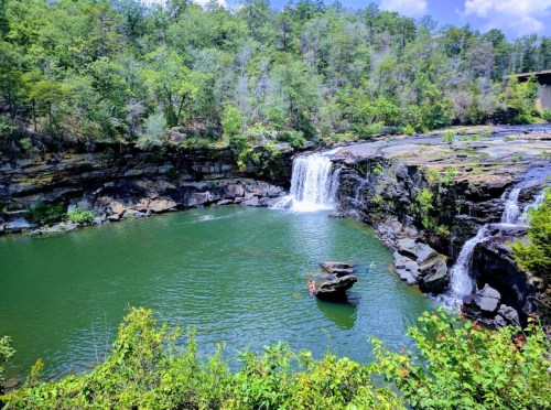 A serene landscape featuring a waterfall cascading into a green pool, surrounded by lush trees and rocky cliffs.