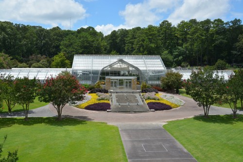 A large glass greenhouse surrounded by colorful flower beds and lush green trees under a blue sky.