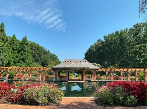 A serene garden with a pond, surrounded by colorful flowers and a gazebo under a clear blue sky.