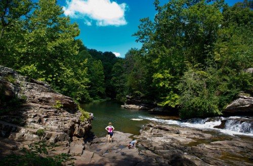 A serene river scene with lush green trees, rocky banks, and two people exploring near the water under a blue sky.