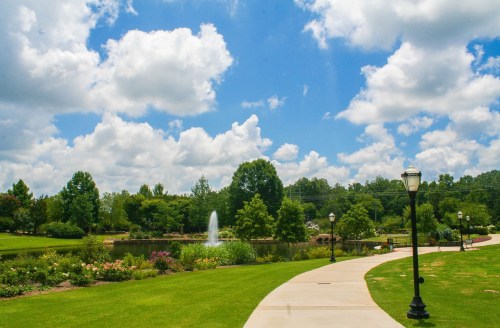 A scenic park pathway lined with lampposts, leading to a pond with a fountain, surrounded by lush greenery and clouds.