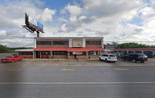 A two-story brick building with a red roof, featuring a sign and parked cars in front under a cloudy sky.