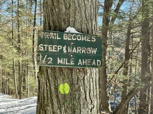 Sign on a tree indicating the trail becomes steep and narrow ½ mile ahead, surrounded by snowy forest.