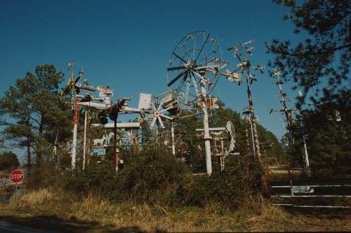A collection of colorful, rusted windmills and sculptures against a clear blue sky, surrounded by trees and grass.