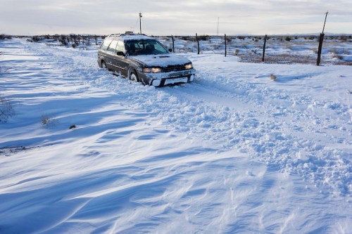 A vehicle stuck in deep snow on a rural road, surrounded by a snowy landscape and a fence in the background.
