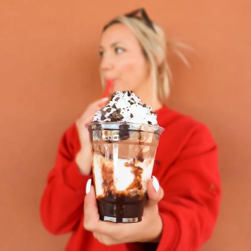 A person in a red sweater holds a dessert cup with whipped cream and chocolate, sipping a drink in the background.