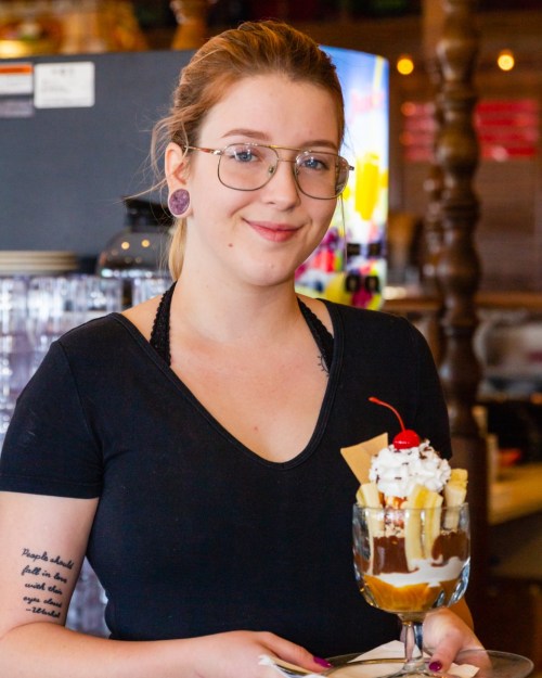 A smiling young woman with glasses holds a dessert topped with whipped cream and a cherry in a café setting.