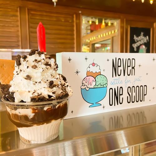 A delicious ice cream sundae topped with whipped cream, chocolate sauce, and a cherry, next to a sign that reads "Never settle for just one scoop."