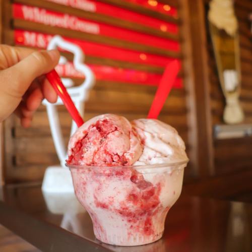 A hand holding a cup of two scoops of strawberry and vanilla ice cream with red spoons, set against a wooden background.