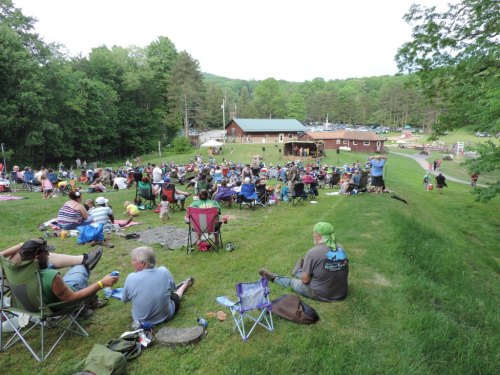 A large crowd gathers on a grassy hill for an outdoor event, with a stage and trees in the background.