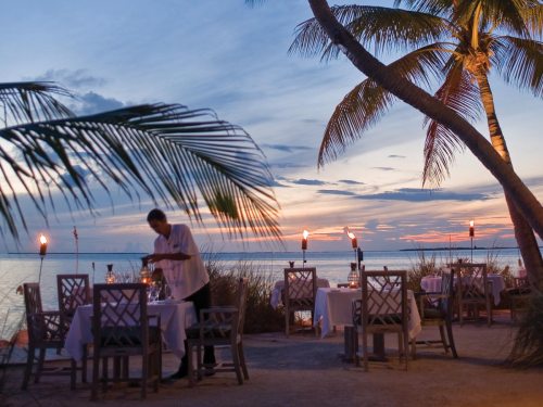 A serene beachside restaurant at sunset, with a waiter setting a table surrounded by palm trees and tiki torches.