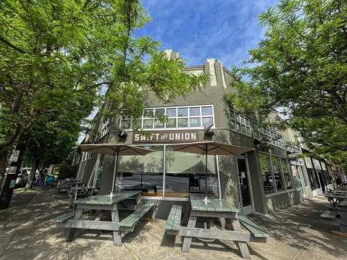 Exterior of Swift and Union restaurant with outdoor seating under umbrellas, surrounded by trees and a blue sky.