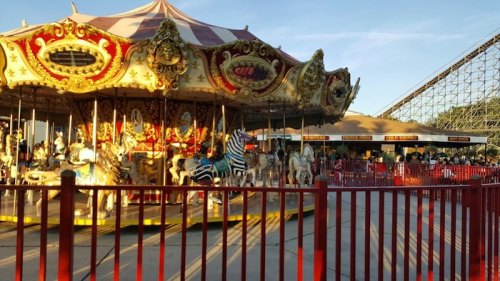 A colorful carousel with ornate decorations, surrounded by a red fence, near a roller coaster in an amusement park.