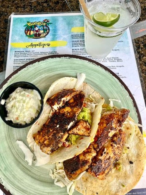 Two fish tacos with guacamole and slaw, served with a side of sauce and a margarita on the rocks. Menu in background.