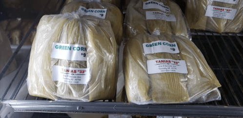 Packages of green corn tamales displayed on a shelf, labeled with prices and store information.
