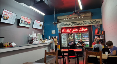 A cozy restaurant interior with a menu board, patrons dining, and a colorful drink fridge in the background.