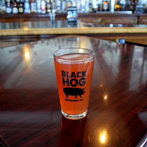 A glass of pink beverage with "BLACK HOG BREWING CO." logo on a wooden bar top, with blurred bottles in the background.