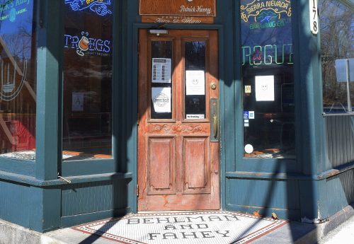 Exterior of a tavern with a wooden door, featuring a quote about American liberty and colorful neon signs.