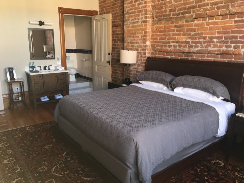 Cozy bedroom with a brick wall, a large bed, nightstands, and an ensuite bathroom visible in the background.