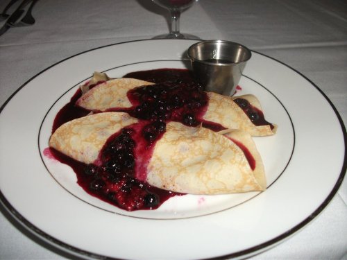 A plate of crepes topped with berry sauce and served with a small metal cup of syrup.
