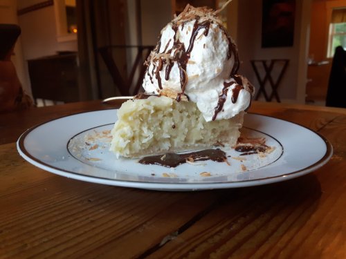 A slice of dessert topped with whipped cream, chocolate drizzle, and coconut flakes on a decorative plate.