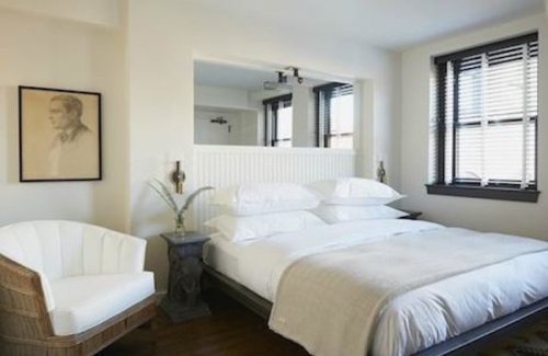 A cozy bedroom featuring a large bed, a white armchair, and a portrait on the wall, with natural light from the windows.