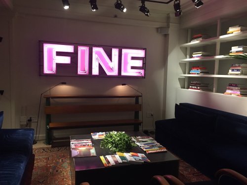 A cozy lounge with a neon "FINE" sign, bookshelves, a coffee table with magazines, and plush seating.