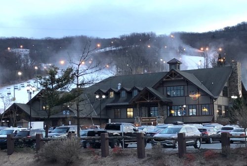 A ski resort lodge with snow-covered slopes and ski lifts in the background, surrounded by parked cars and trees.