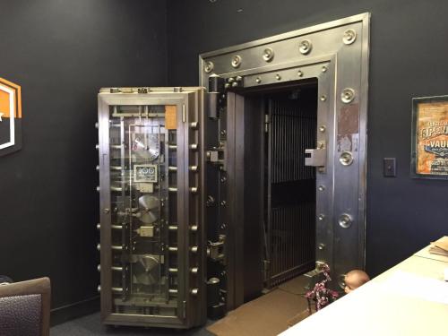 A large, metallic bank vault door stands open next to a smaller, glass-fronted safe in a dimly lit room.