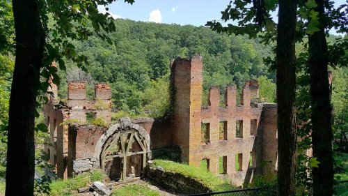 Ruins of a brick building surrounded by trees and greenery, with a clear blue sky in the background.