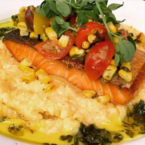 Grilled salmon on creamy grits, topped with cherry tomatoes, corn, and fresh greens, drizzled with sauce.