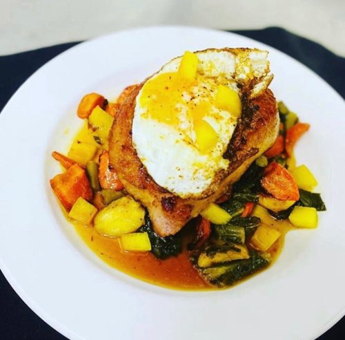 A plate of sautéed vegetables topped with a seared piece of meat and a fried egg, garnished with mango.