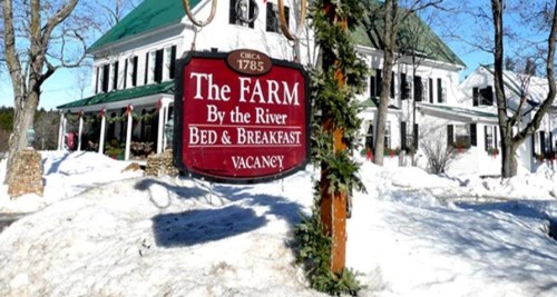 Sign for "The Farm By the River Bed & Breakfast" with snow-covered ground and a historic building in the background.