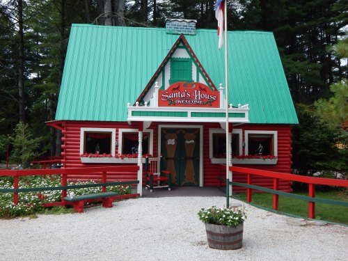 A charming red and green cabin with a sign reading "Santa's House," surrounded by trees and flowers.