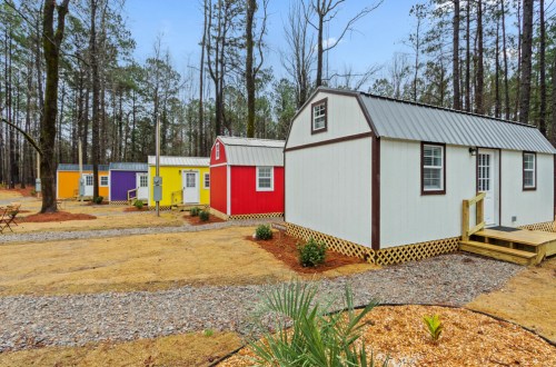 Colorful tiny cabins in a wooded area, surrounded by gravel paths and greenery.