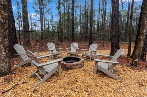 A cozy fire pit area with six wooden chairs surrounded by gravel and trees in a serene outdoor setting.