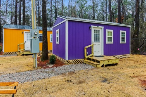 Two colorful cabins, one purple and one orange, surrounded by gravel and pine trees. A utility pole is visible nearby.