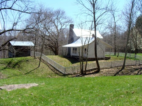 A white house with a porch and a chimney, surrounded by trees and a white fence, on a sunny day.