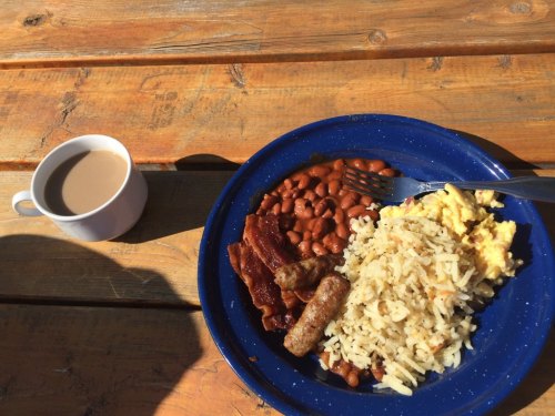 A blue plate with scrambled eggs, sausage, bacon, baked beans, and hash browns, alongside a cup of coffee on a wooden table.
