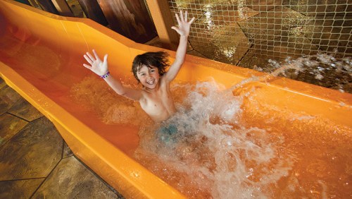 A joyful child splashes into the water at the end of an orange waterslide, arms raised in excitement.