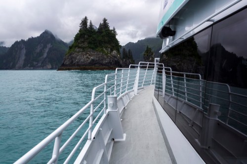 A boat's railing curves along the side, with rocky islands and misty mountains in the background on a cloudy day.
