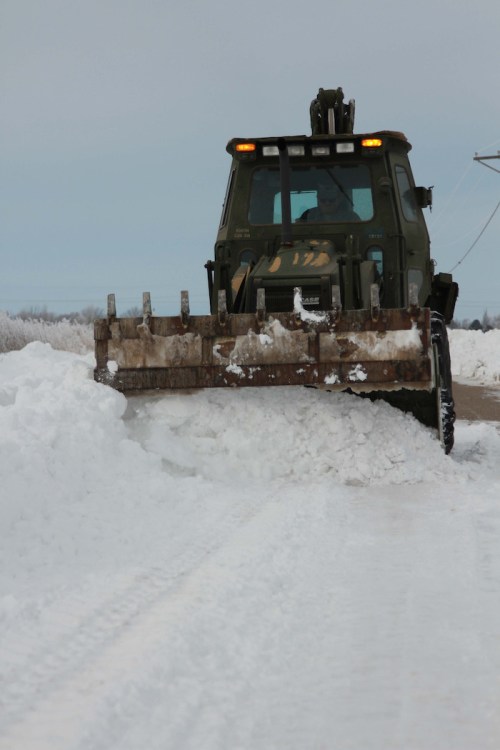 A snowplow clears a snowy road, with large piles of snow on either side and a cloudy sky above.