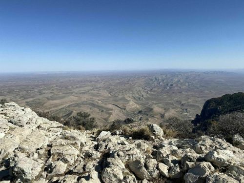 A panoramic view of a vast, arid landscape from a rocky vantage point under a clear blue sky.