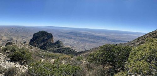 A panoramic view of a mountainous landscape with a clear blue sky and distant valleys. Green shrubs in the foreground.