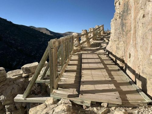 A wooden bridge on a rocky cliff, with shadows cast by the railing, overlooking a mountainous landscape.