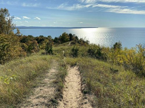A sandy path leads through grassy hills to a shimmering lake under a clear blue sky with scattered clouds.