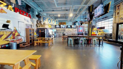 A spacious food hall with colorful signage, tables, and a bar area, featuring various food vendors and a modern design.