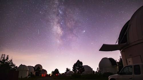 A starry night sky with the Milky Way above observatories and trees, illuminated by soft lights.