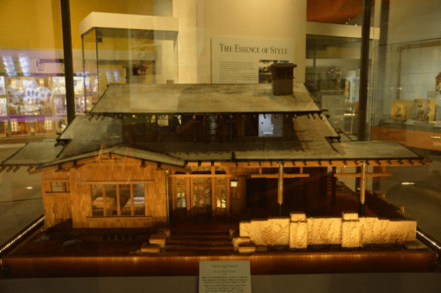 A detailed model of a wooden house with a sloped roof, displayed in a glass case at a museum.