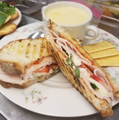 A grilled sandwich with turkey, tomatoes, and spinach, served with a cup of soup and crackers on a floral plate.
